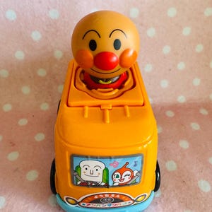 May include: An orange toy bus with a cartoon character on top, featuring a round head, red nose, and rosy cheeks. The bus has black wheels, a blue base, and a license plate that reads "A-123". The bus has a cartoon image on the side.