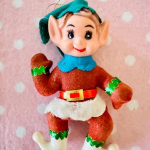 May include: A vintage Christmas elf ornament. The elf has a red body, white boots, and a white skirt. It has a blue collar and hat, with green accents. The elf has a red belt with a gold buckle. The ornament is on a pink background.