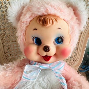 May include: A vintage-style plush doll with a pink bear-like head and a fluffy pink body. The doll has blue eyes, rosy cheeks, and a red mouth. It wears a pink hat with white trim and a striped ribbon bow. The doll is seated in a wicker chair.