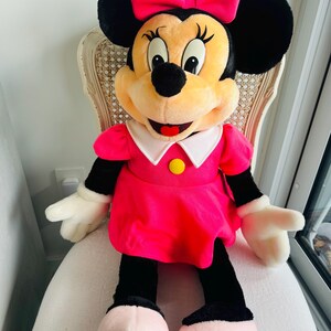 Może przedstawiać: Duża pluszowa zabawka Minnie Mouse w r&oacute;żowej sukience i r&oacute;żowej kokardce. Ma czarne uszy, czarny nos i szeroki uśmiech.