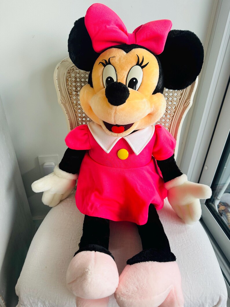 Może przedstawiać: Duża pluszowa zabawka Minnie Mouse w r&oacute;żowej sukience i r&oacute;żowej kokardce. Ma czarne uszy, czarny nos i szeroki uśmiech.
