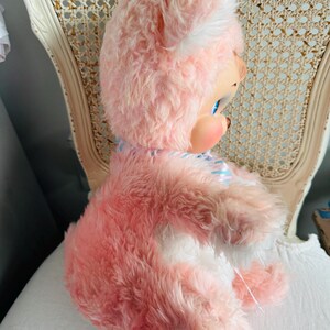 May include: A vintage pink plush bear doll with a white face and ears, blue eyes, and rosy cheeks. The doll has a blue and white striped bow and is seated on a white surface. The bear is a soft, plush toy.
