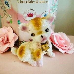May include: A white and gold ceramic figurine of a lamb with a bell around its neck. The lamb is sitting on a pink surface with pink flowers.
