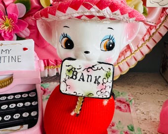 May include: A white ceramic piggy bank wearing a red and white striped dress and a pink straw hat. The piggy bank is holding a sign that says "Bank".