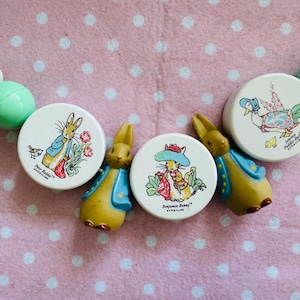 Può includere: Un giocattolo per bambini a tema Peter Rabbit con illustrazioni rotonde e bianche di Peter Rabbit e Benjamin Bunny, insieme a un'anatra. Il giocattolo ha figure di coniglietti gialli e blu e perline verdi e bianche. Lo sfondo è un tessuto rosa con pois bianchi.