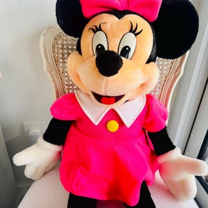Może przedstawiać: Duża pluszowa zabawka przedstawiająca Minnie Mouse w r&oacute;żowej sukience z białym kołnierzykiem i ż&oacute;łtym guzikiem. Ma r&oacute;żową kokardkę we włosach i szeroki uśmiech.