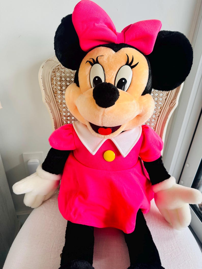 Może przedstawiać: Duża pluszowa zabawka przedstawiająca Minnie Mouse w r&oacute;żowej sukience z białym kołnierzykiem i ż&oacute;łtym guzikiem. Ma r&oacute;żową kokardkę we włosach i szeroki uśmiech.