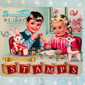 May include: A vintage-style tin box with a blue background and a colourful illustration of two children playing with alphabet blocks. The box features the text "Dumpling Dynasty Making Life Sweeter" and "Alphabet Stamps".