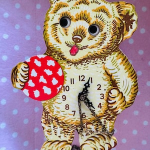 May include: A vintage bear-shaped clock with a red and white polka-dot ball. The clock face has black numerals and hands, with a chain pendulum. The bear is beige and brown, with googly eyes and a pink tongue. Made in Germany.
