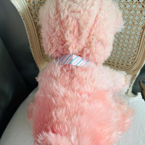 May include: A plush pink stuffed animal with a fluffy texture. The toy has a light blue and pink striped collar. The toy is sitting on a white surface and a vintage chair.