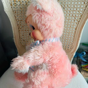May include: A vintage pink plush toy bunny with a white and blue striped collar. The bunny has a round face with painted features and long, fluffy ears. It is sitting on a white chair.