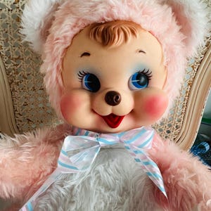 May include: A vintage plush doll with a pink fur hat and matching fur body. The doll has a smiling face with blue eyes, rosy cheeks, and a red mouth. A striped ribbon bow adorns the neck. The doll is seated in a wicker chair.