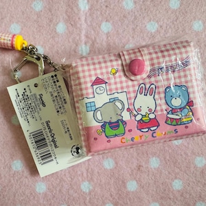 Puede incluir: Una cartera a cuadros rosa y blanca con cierre de cremallera y un llavero adjunto. La cartera presenta una imagen de dibujos animados de una escuela con un oso, un conejo y un elefante tocando música. El texto "Cherry Chums" está impreso en la parte delantera de la cartera. El llavero es un lápiz amarillo y plateado.