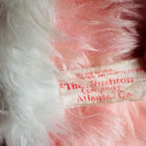 May include: Close-up of a plush toy with pink and white fur. A tag reads "The Rushton Company, Atlanta, Ga.". The toy's fur is soft and fluffy, with a mix of light pink and white colors. The tag is white with red text.