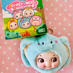 Może przedstawiać: Pluszowy niebieski brelok do kluczy z motywem słonia z uśmiechniętą twarzą lalki. Brelok znajduje się obok pudełka z napisem "PET PET ANGEL COLLECTION" i ilustracjami postaci w kostiumach zwierząt. Pudełko ma napis "FUN RECORD".