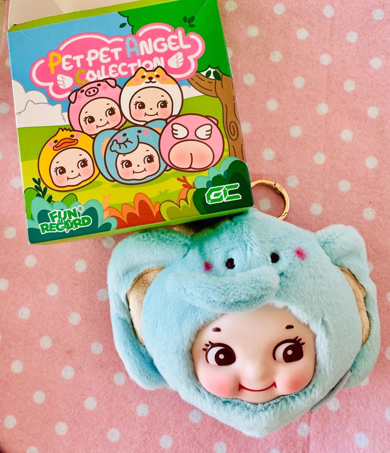 Może przedstawiać: Pluszowy niebieski brelok do kluczy z motywem słonia z uśmiechniętą twarzą lalki. Brelok znajduje się obok pudełka z napisem "PET PET ANGEL COLLECTION" i ilustracjami postaci w kostiumach zwierząt. Pudełko ma napis "FUN RECORD".