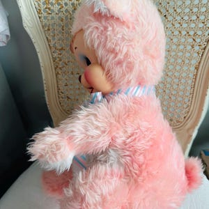 May include: A vintage pink plush bunny doll with a painted face, blue eyes, and a striped ribbon around its neck. The doll has a fluffy, textured appearance and a small, rounded tail. It is sitting on a white surface.
