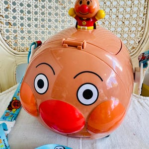 May include: A tan-coloured Anpanman-themed container with a red nose and large black eyes. A small Anpanman figure sits on top. The container has a blue strap with cartoon characters.