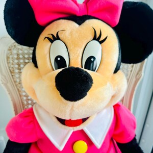 Może przedstawiać: Pluszowa zabawka przedstawiająca Minnie Mouse w r&oacute;żowej sukience z białym kołnierzykiem i ż&oacute;łtym guzikiem. Ma dużą r&oacute;żową kokardkę we włosach i charakterystyczne czarne uszy.