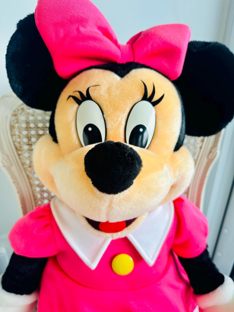 Może przedstawiać: Pluszowa zabawka przedstawiająca Minnie Mouse w r&oacute;żowej sukience z białym kołnierzykiem i ż&oacute;łtym guzikiem. Ma dużą r&oacute;żową kokardkę we włosach i charakterystyczne czarne uszy.