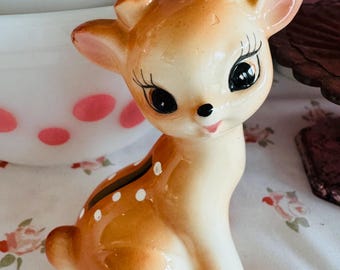 May include: A vintage ceramic deer figurine with large black eyes, long eyelashes, and a small pink mouth. The deer has a light brown coat with white spots and red hooves. The figurine is sitting on a floral patterned surface.