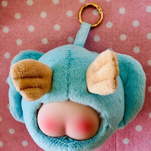 Może przedstawiać: Niebieski pluszowy brelok w kształcie postaci z kresk&oacute;wki, z brązowymi uszami i r&oacute;żowym tyłkiem. Posiada złoty pierścień i niebieską metkę z napisem "IN RECORD". Tło jest r&oacute;żowe z białymi kropkami.