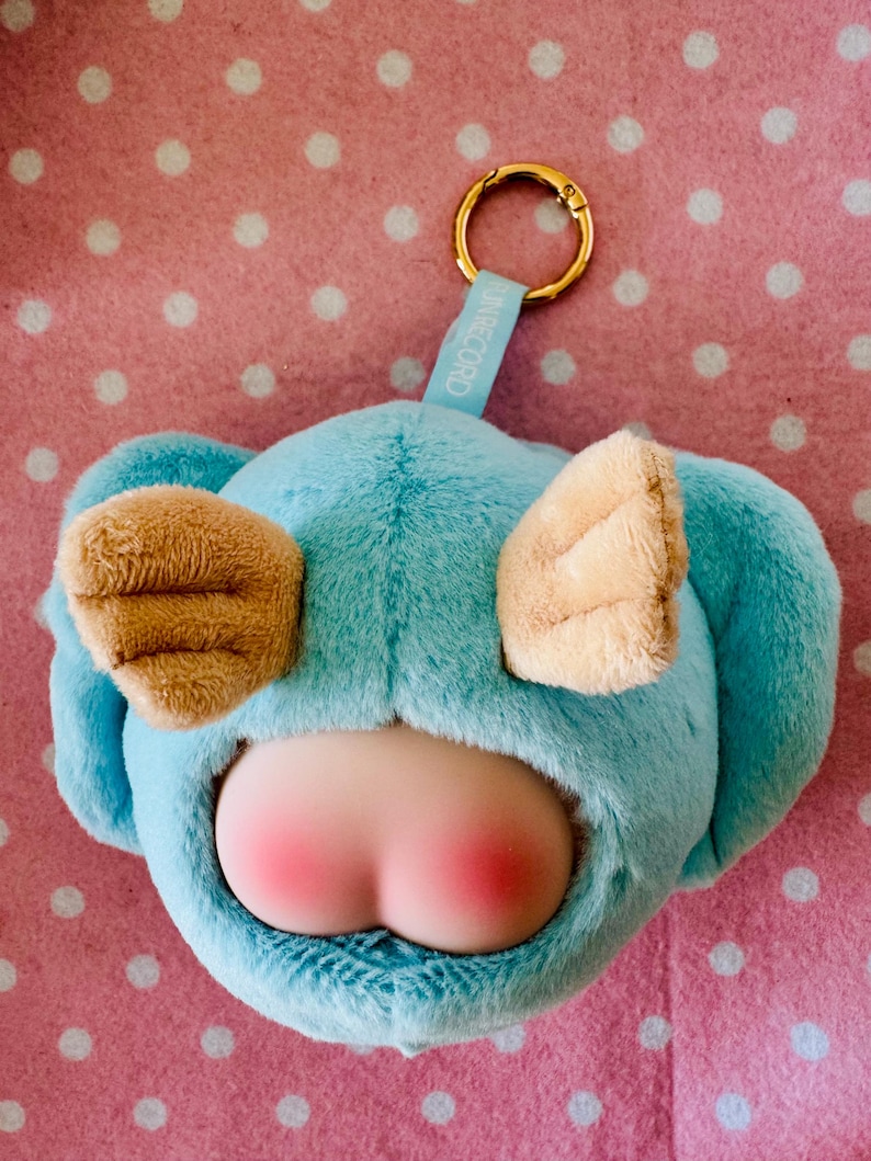 Może przedstawiać: Niebieski pluszowy brelok w kształcie postaci z kresk&oacute;wki, z brązowymi uszami i r&oacute;żowym tyłkiem. Posiada złoty pierścień i niebieską metkę z napisem "IN RECORD". Tło jest r&oacute;żowe z białymi kropkami.