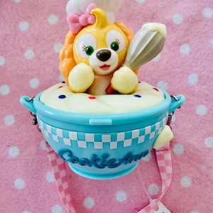 May include: A yellow cartoon dog figure wearing a chef's hat and pink bow, holding a whisk, emerging from a light blue bowl filled with cream. The bowl has the text "Cookie Ann" and a pink and white checkered strap.