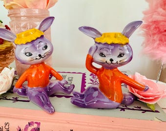 May include: Two purple ceramic bunny figurines with orange clothing and yellow headbands. The bunnies are sitting on a book with a pink cover.