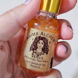May include: A small glass bottle of Anima Alchemia Venus Ritual Oil. The bottle has a gold cap and a white dropper. The label features a woman's portrait and the words "Venus, Attraction, Opulence, and Sensual Joy". The oil inside is a shimmering gold color.
