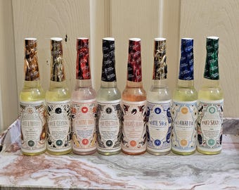 May include: Eight glass bottles with colorful labels and foil-wrapped tops are arranged on a decorative tray. The labels feature text such as "House Cleansing", "Protection", and "Palo Santo". The bottles contain a yellow liquid.