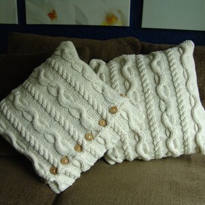 May include: Two white knitted throw pillows with a cable knit pattern and wooden buttons. The pillows are on a brown couch.