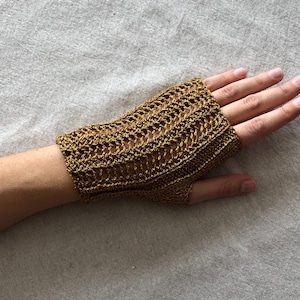 May include: A gold-colored, open-weave fingerless glove. The glove is made of a metallic thread and has a textured appearance. The wrist area is solid, while the hand portion has a decorative, open pattern. The glove is worn on a person's hand.