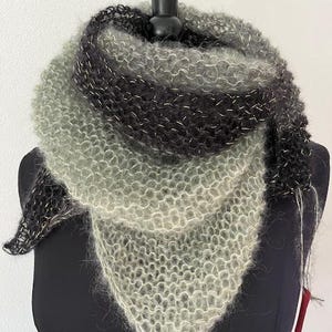 May include: A hand-knitted triangular shawl in shades of gray, black, and silver. The shawl is displayed on a black mannequin and features a textured, open-weave design. The color gradient transitions from light gray to black, with silver threads woven throughout.