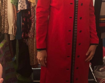 Puede incluir: Una mujer lleva un abrigo rojo con ribetes negros y un vestido a rayas blancas y negras. El abrigo tiene cuello y botones en la parte delantera.
