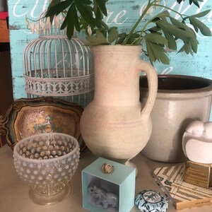 May include: A still life featuring a white ceramic jug with a handle, a white birdcage, a white ceramic elephant figurine in a light blue box, a white glass bowl with a textured surface, a small round tin with a blue and white floral design, and a book with the text "How to Wear" on the cover.