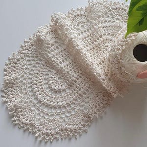 Może przedstawiać: Ręcznie robiona serwetka szydełkowa w kolorze kremowym, o okrągłym wzorze i ząbkowanych krawędziach. Widoczna jest kulka białej przędzy i różowy szydełko, co sugeruje projekt rzemieślniczy. Serwetka wykonana jest z miękkiej, teksturowanej przędzy.