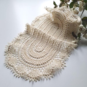 Puede incluir: Camino de mesa de ganchillo hecho a mano, de color crema, con un diseño circular y bordes festoneados. La delicada textura de encaje es visible, con una sección enrollada en la parte superior. Una ramita de bayas blancas y hojas verdes está en el fondo.