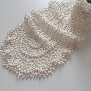 Puede incluir: Tapete de ganchillo hecho a mano, de color crema, con un borde festoneado. El delicado diseño calado está drapeado sobre una superficie, con un ovillo de hilo a juego visible. El tapete presenta un patrón circular.
