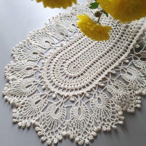 Puede incluir: Tapete de ganchillo color crema, intrincadamente elaborado con un borde festoneado. El encaje presenta un diseño calado detallado con patrones de lágrima y circulares. Flores amarillas brillantes en el fondo.