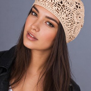 May include: A beige crocheted hat with an openwork design. The hat is made of a light-colored yarn and has a rounded shape. The hat is worn on the head. The background is a solid gray color.