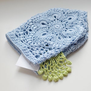 May include: A light blue crocheted hat with a detailed floral pattern. A small, folded white tag is tucked inside, and a green fringe detail is visible. The hat is made of soft, textured yarn.