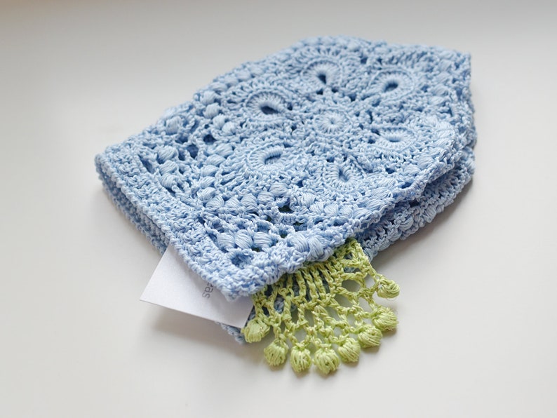 May include: A light blue crocheted hat with a detailed floral pattern. A small, folded white tag is tucked inside, and a green fringe detail is visible. The hat is made of soft, textured yarn.