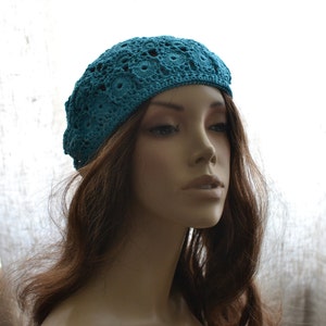 May include: A teal blue crocheted beret with a delicate openwork pattern. The beret is worn on a mannequin with long brown hair.