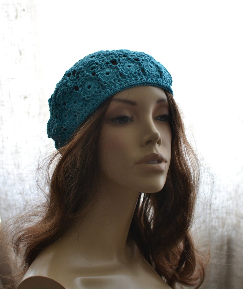 May include: A teal blue crocheted beret with a delicate openwork pattern. The beret is worn on a mannequin with long brown hair.