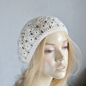 May include: A white crocheted beret with a delicate openwork pattern. The beret is worn on a mannequin with blonde hair.