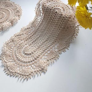 Puede incluir: Dos tapetes beige tejidos a ganchillo sobre una superficie blanca. Uno es redondo, el otro tiene una forma larga y curvada. El tejido de ganchillo presenta patrones intrincados y un delicado borde con flecos. Flores amarillas en el fondo.