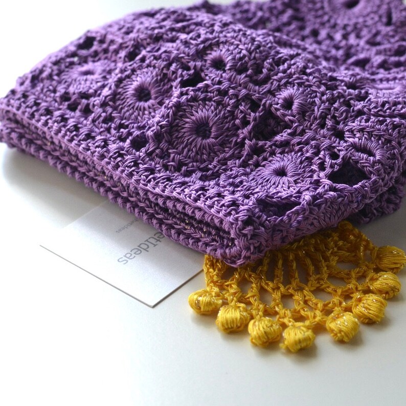 May include: A purple crocheted shawl with a yellow crocheted fringe. The shawl is folded and the fringe is visible on the bottom edge. The shawl is on a white surface with a white card that says "eludes".