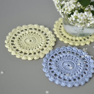 Op de afbeelding: Drie handgemaakte gehaakte onderzetters in pastelkleuren. Eén is lichtgeel, een andere lichtblauw, en de derde is ook lichtgeel met een glas water en witte bloemen op de achtergrond. De onderzetters zijn cirkelvormig met een gedetailleerd ontwerp.