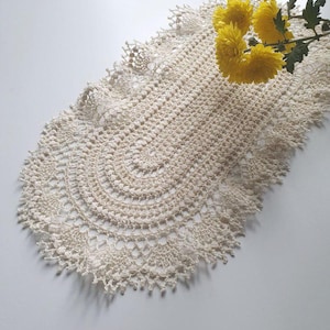 Puede incluir: Camino de mesa de ganchillo de color crema, de forma ovalada, con un diseño calado detallado. El camino de mesa presenta un borde festoneado y está adornado con flores de crisantemo amarillas. El textil hecho a mano añade un toque de encanto vintage.